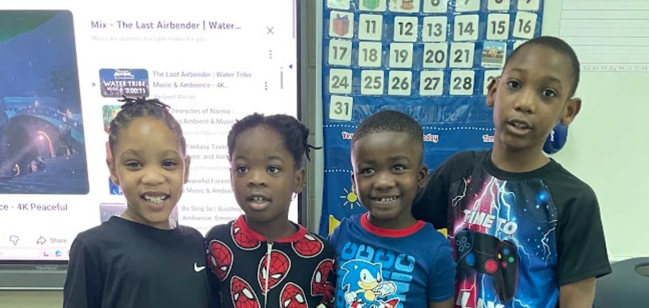 Four children stand close together in front of a white board with a calendar and a monitor showing a website.