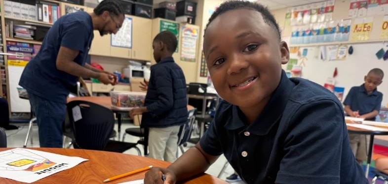 A child wearing a blue shirt smiles while sitting at a desk and holding a pencil.