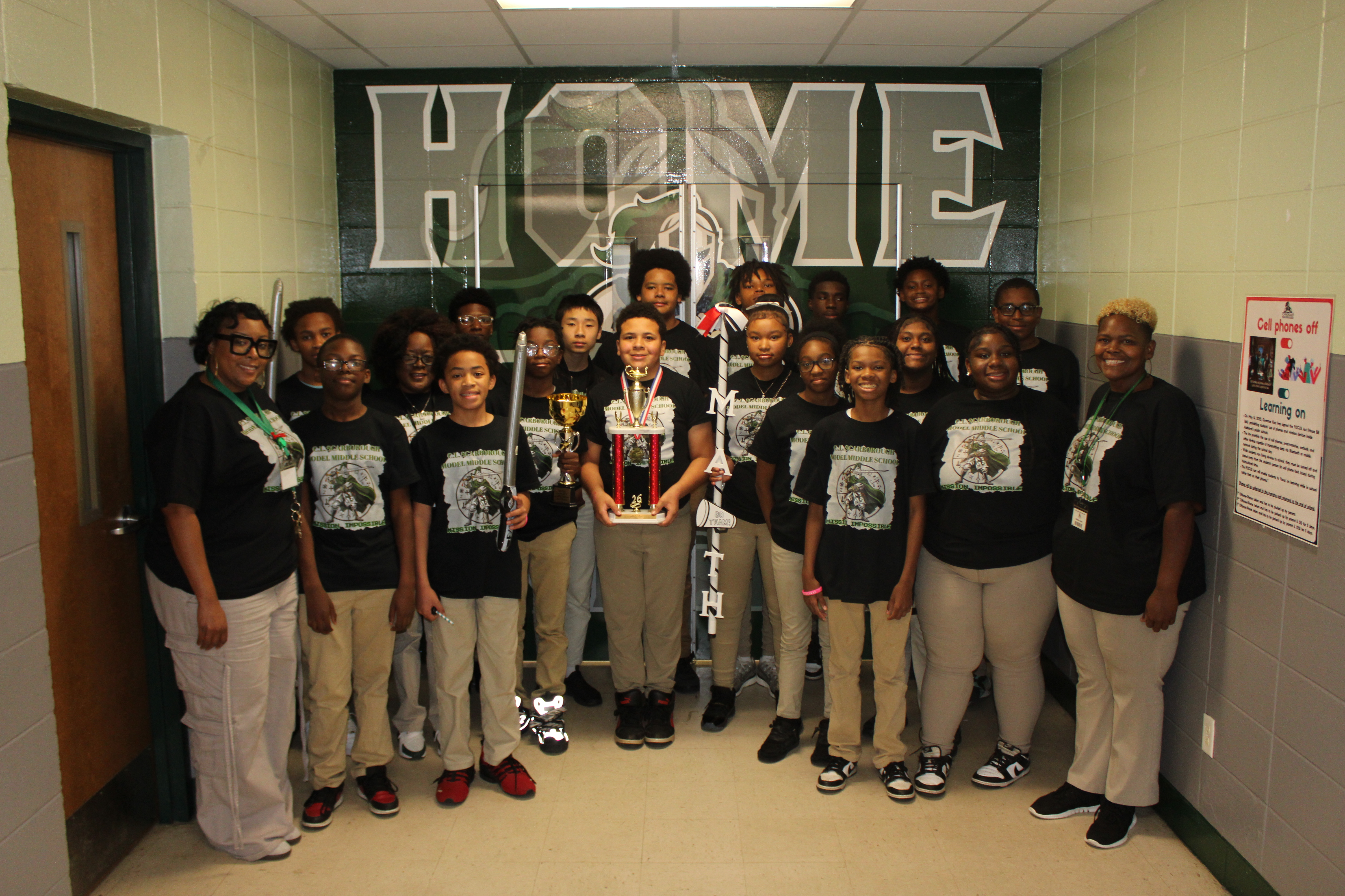 A group of people in matching black T-shirts pose for a photo, with a wall in the background displaying the word "HOME".