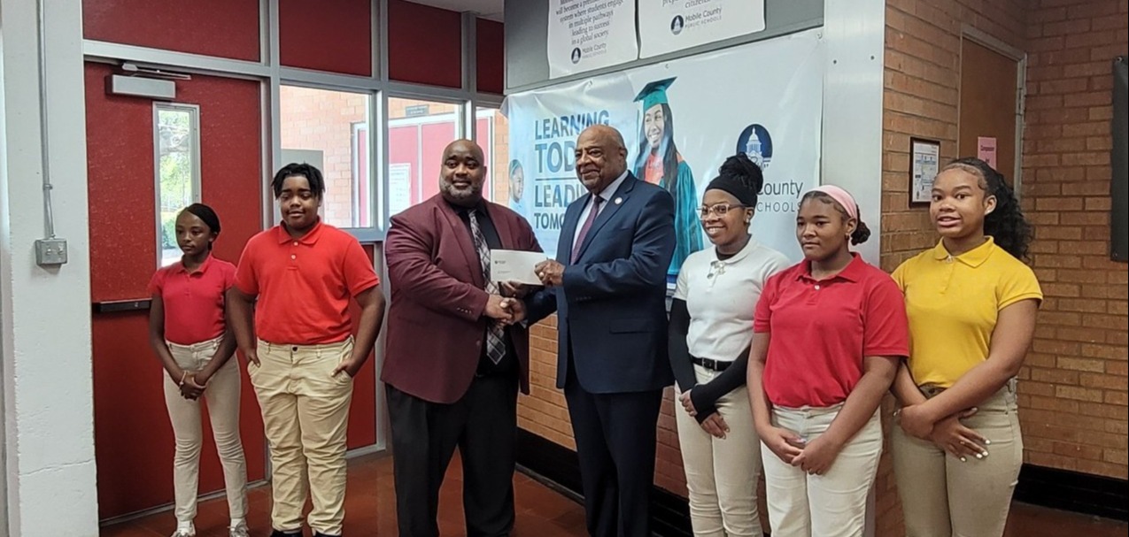 Students standing around Mr. Williams and Sam Jones while Sam Jones present Mr. Williams with a check.
