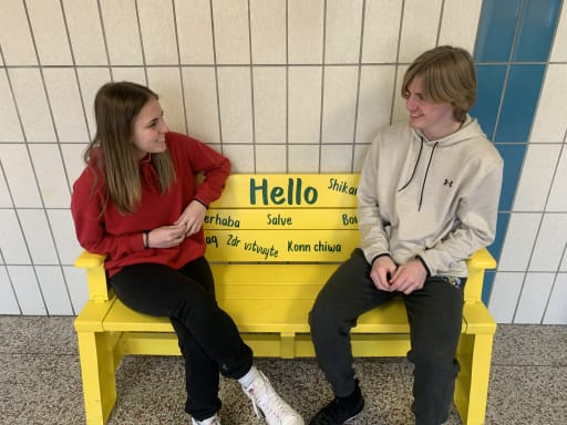 Two teenagers are sitting on a bright yellow bench with the word 'Hello' and various greetings in different languages painted on it, engaged in conversation in a school hallway.