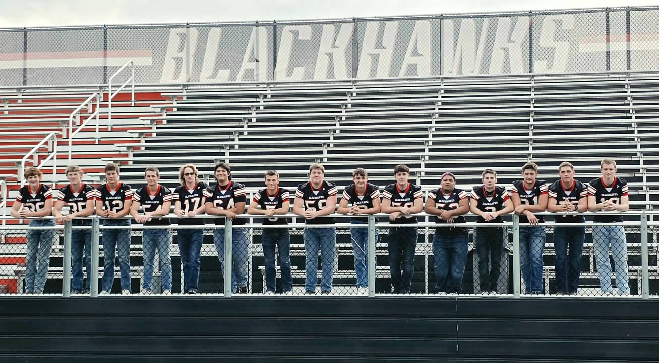 Senior football players lined up along fence on bleachers.