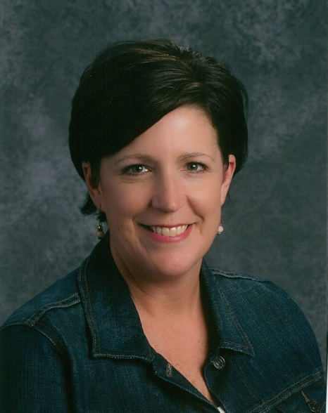 A woman wearing a denim jacket over a white shirt, standing confidently with a neutral background.
