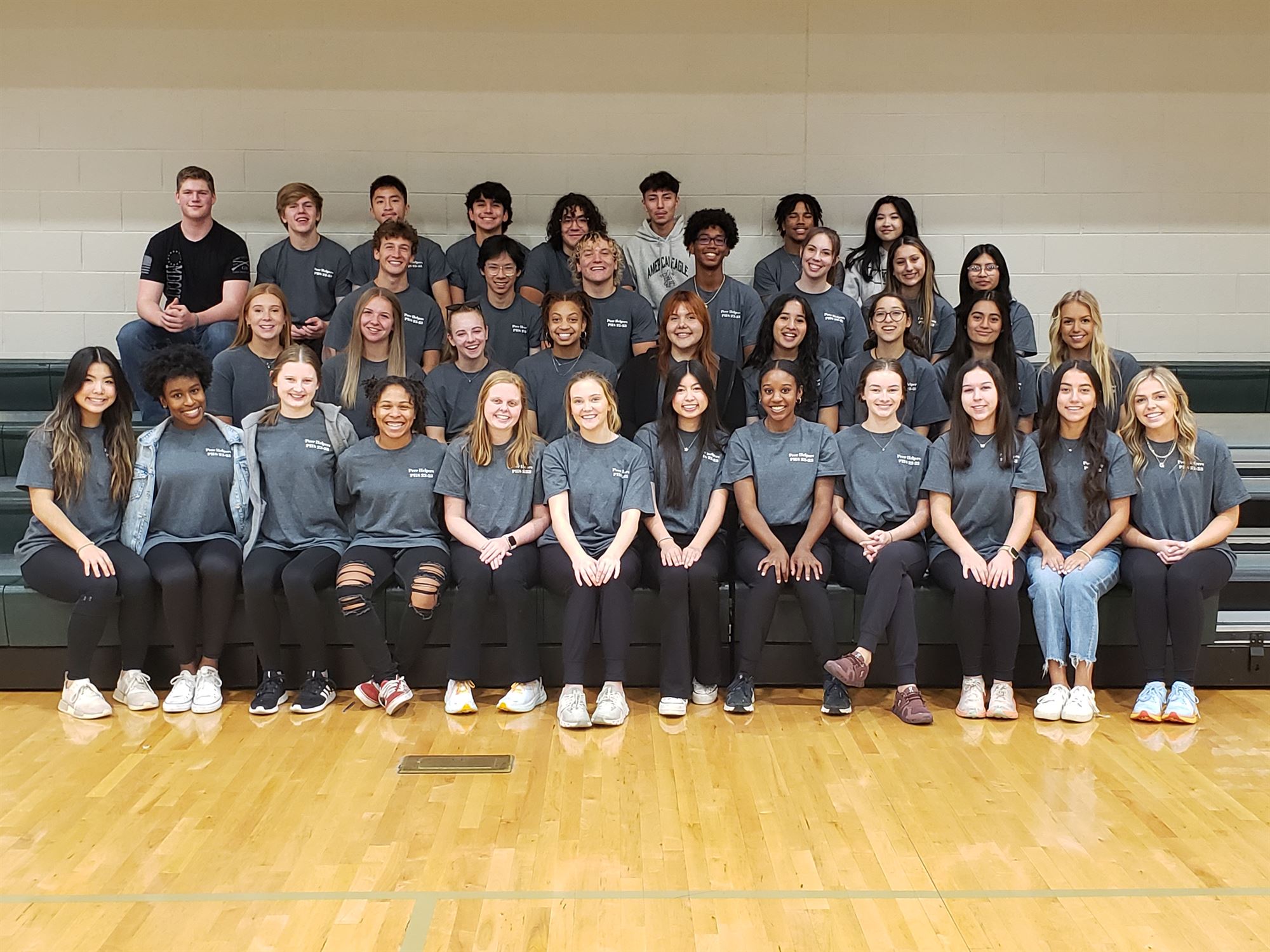 1. A group of young individuals in black shirts seated together on a bench, engaged in conversation and enjoying each other's company. 2. Young people wearing black shirts are sitting on a bench, sharing a moment of camaraderie and laughter in a casual setting. 3. A diverse group of young adults in black shirts sits on a bench, creating a lively atmosphere as they interact with one another.