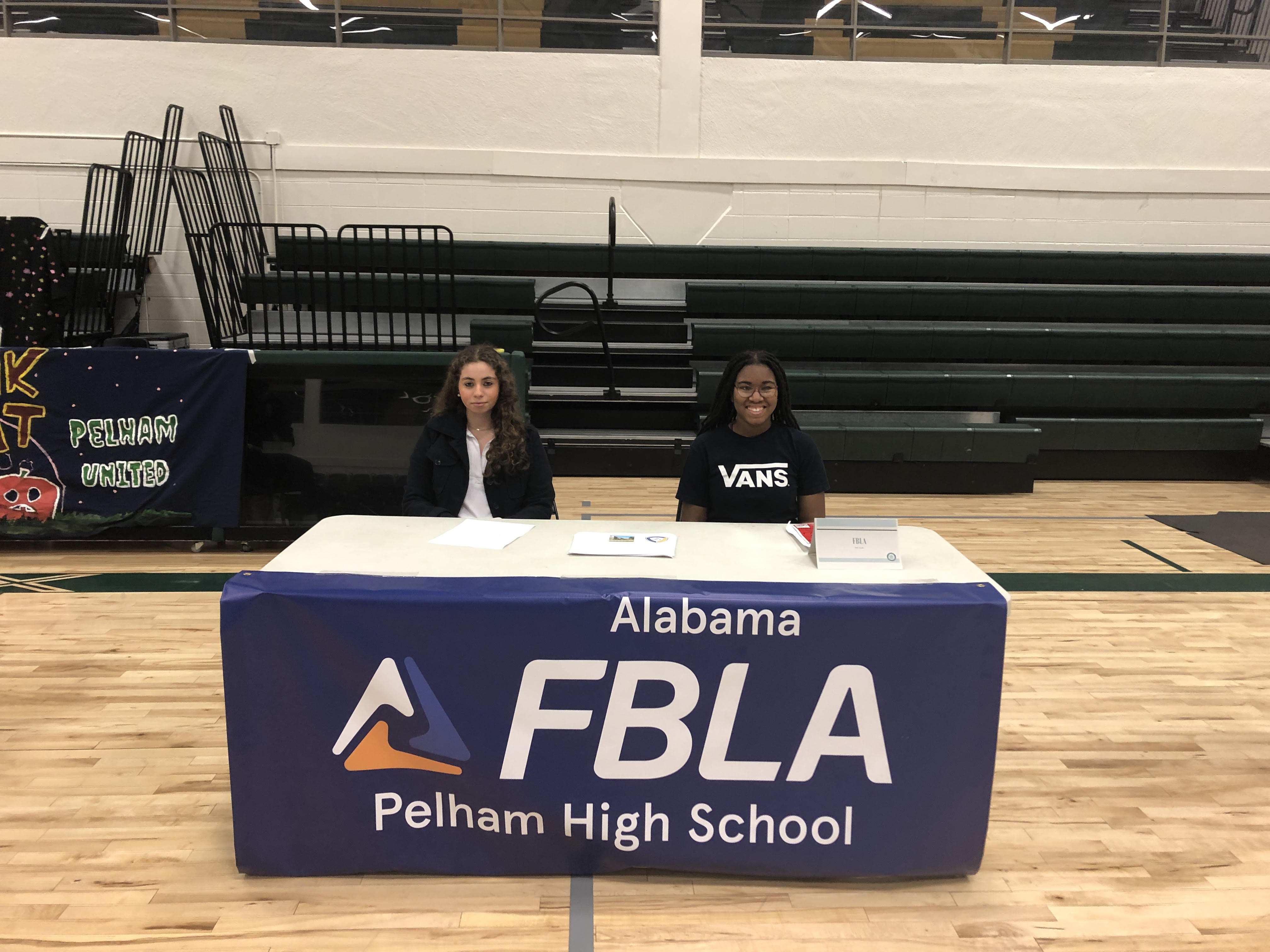 Two students sit behind a table covered with a blue banner that reads "Alabama FBLA Pelham High School" in a school gymnasium. The student on the left has long curly hair and is wearing a black jacket over a white shirt, while the student on the right is smiling and wearing a black "VANS" T-shirt. Papers and a small nameplate labeled "FBLA" are on the table. Behind them, green bleachers and a decorated banner with "Pelham United" can be seen.