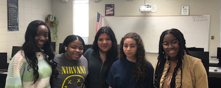 Five young women smile and pose together for a photo in a bright classroom setting.