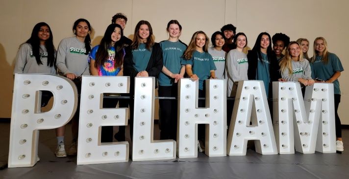 1. A diverse group of young individuals smiling and posing in front of a sign that reads "Pelham."  2. Young people of various backgrounds standing together, smiling in front of a sign labeled "Pelham."  3. A cheerful group of young adults posing in front of a sign that displays the name "Pelham."