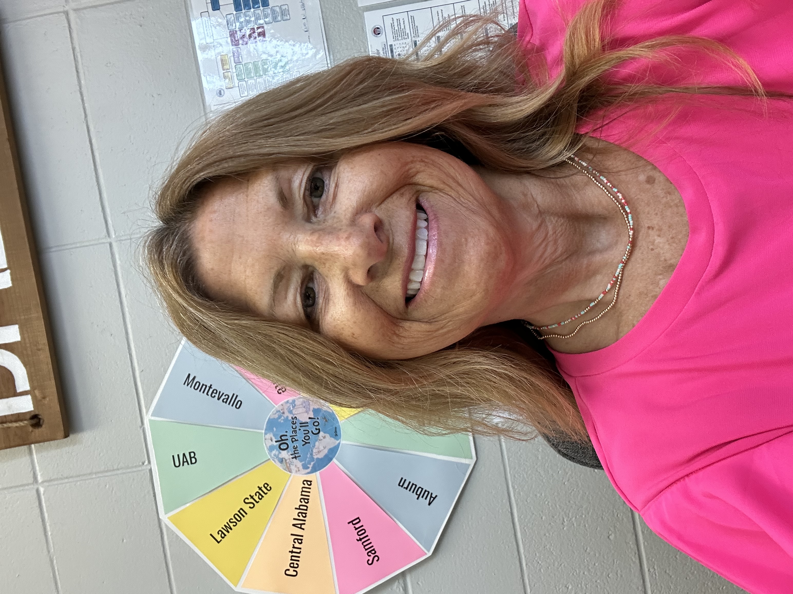 A woman in a pink shirt smiles brightly in front of a large wheel, exuding joy and positivity.