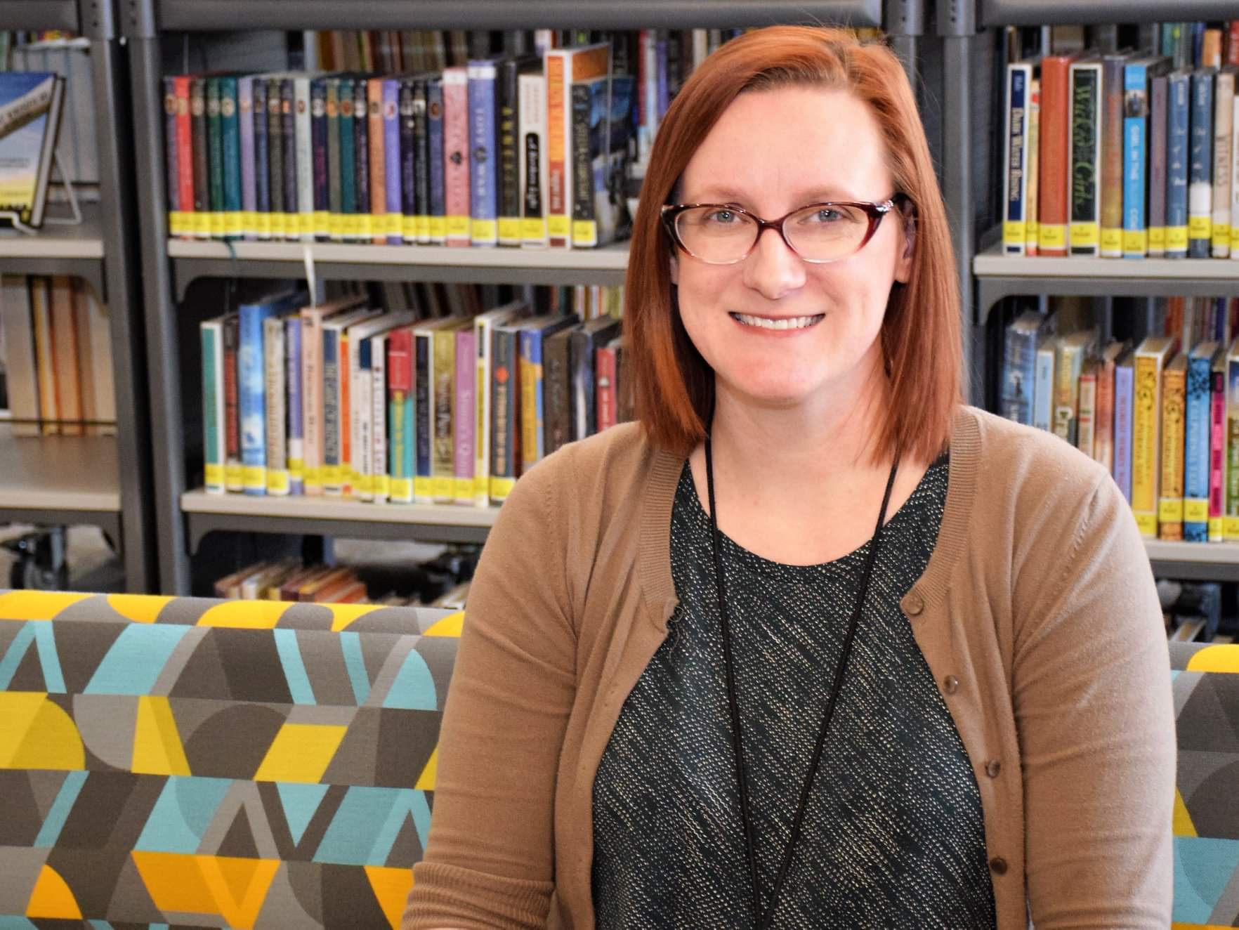 A woman seated on a couch, surrounded by bookshelves filled with books in a cozy library setting.