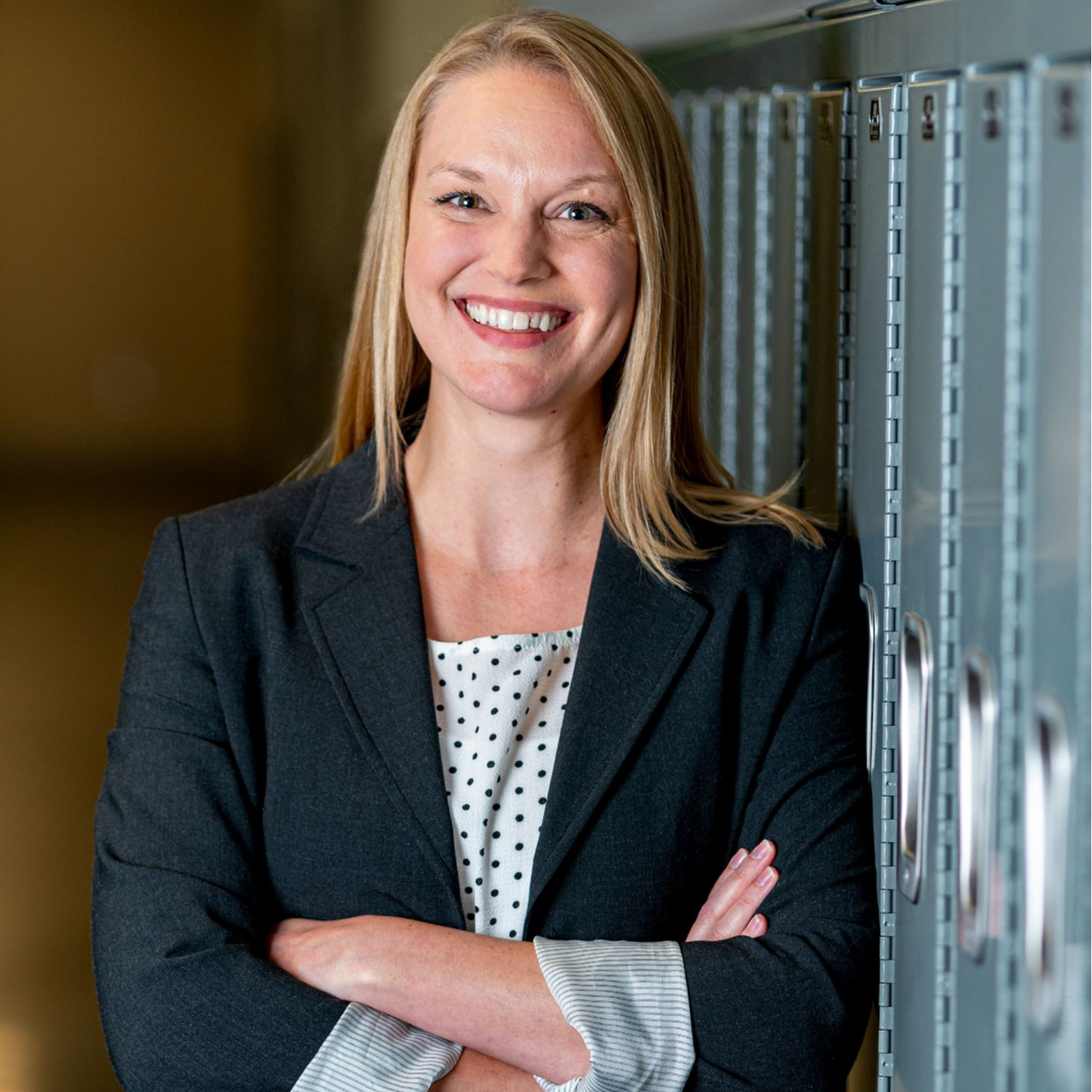A professional woman in a business suit stands confidently beside a row of lockers in a workplace setting.