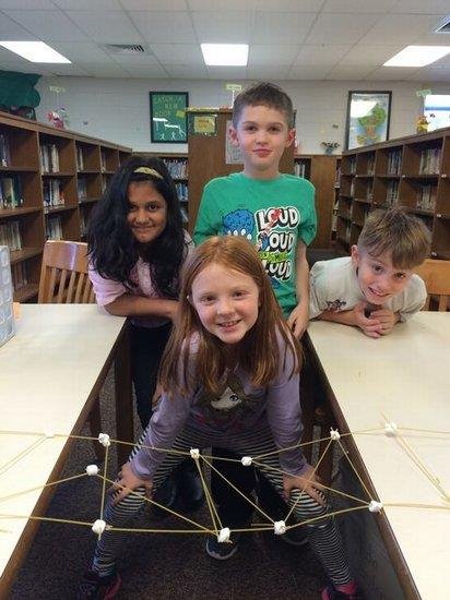 Three children smile for a photo in front of a tall bookcase filled with colorful books.