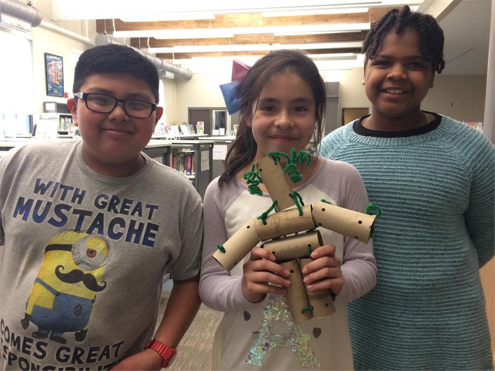Three children smile for a photo, standing beside a colorful cardboard robot they created together.