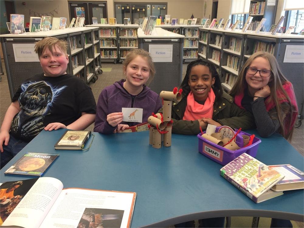 Four girls seated at a table, engaged with books and a single book in front of them, sharing a moment of study.