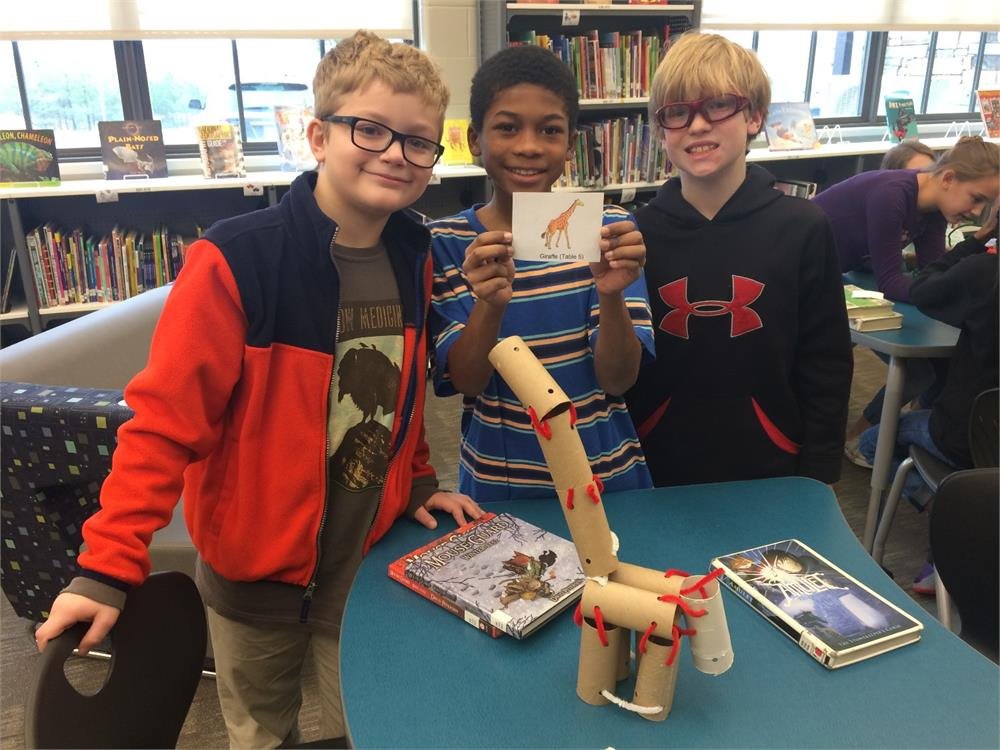 Three children smile for a photo, standing beside a large cardboard giraffe in a cheerful setting.