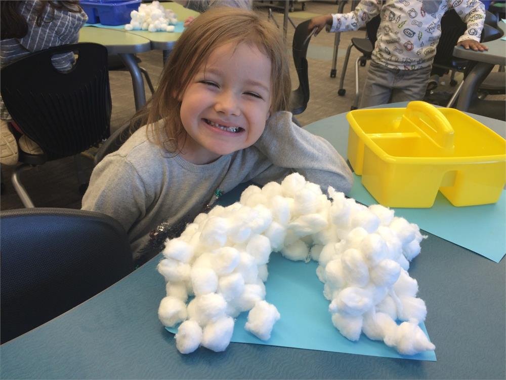 A young girl smiles joyfully at a table, with a fluffy cloud floating nearby, creating a whimsical atmosphere.