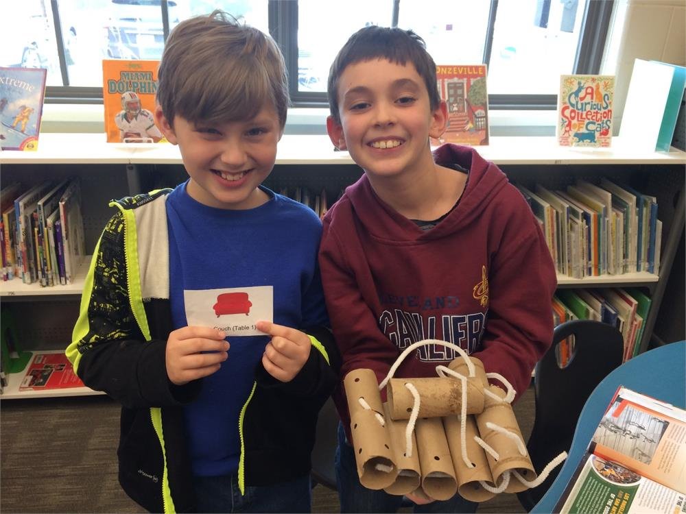 Two boys proudly display cardboard tubes in front of a colorful bookcase filled with various books.