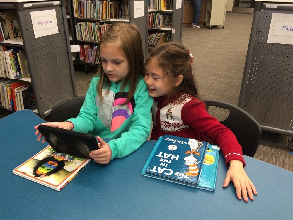 Two girls seated at a table, engaged with books and a tablet, showcasing a moment of study and collaboration.