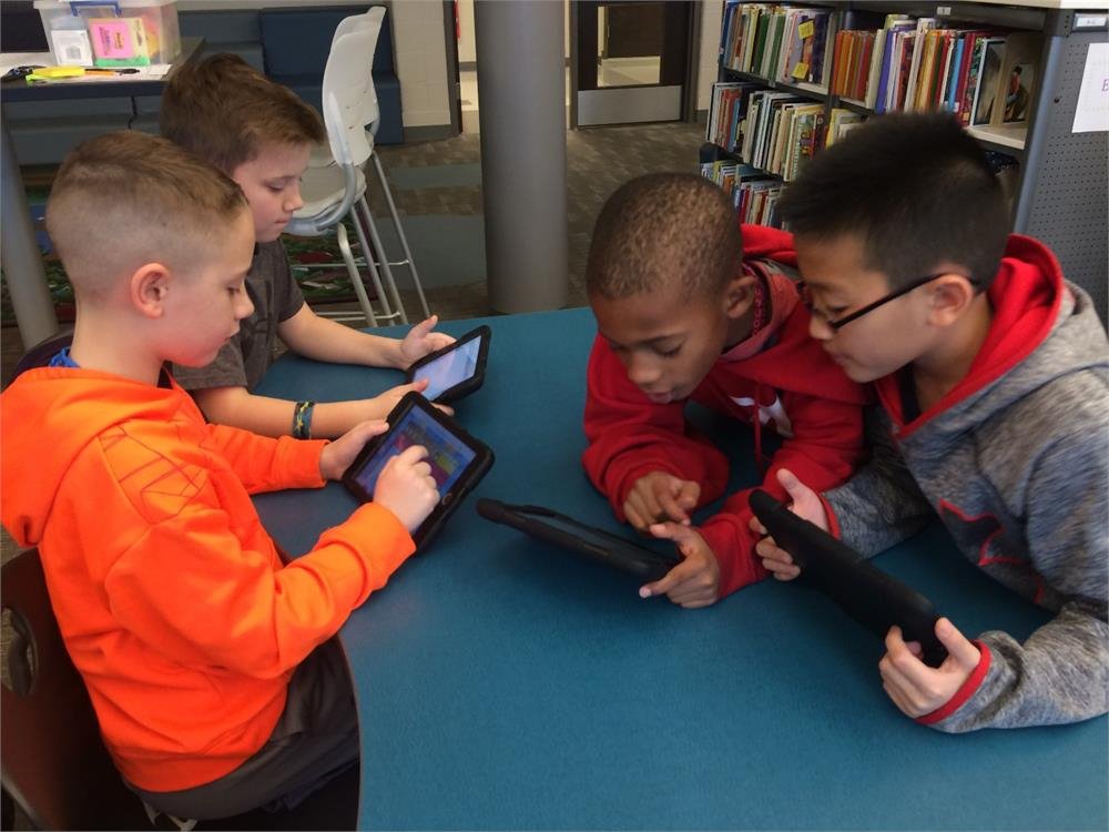 Four children engaged with their tablets while sitting around a table, focused on their screens and sharing ideas.