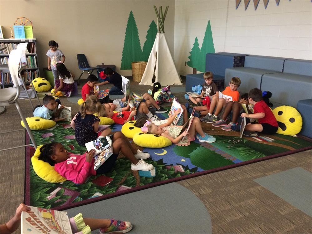 Children seated on colorful bean bags in a library, engrossed in reading books, surrounded by shelves of literature.