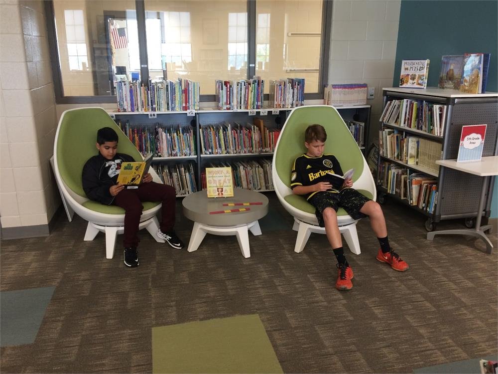 Two boys seated in chairs, engaged in reading within a quiet library setting.