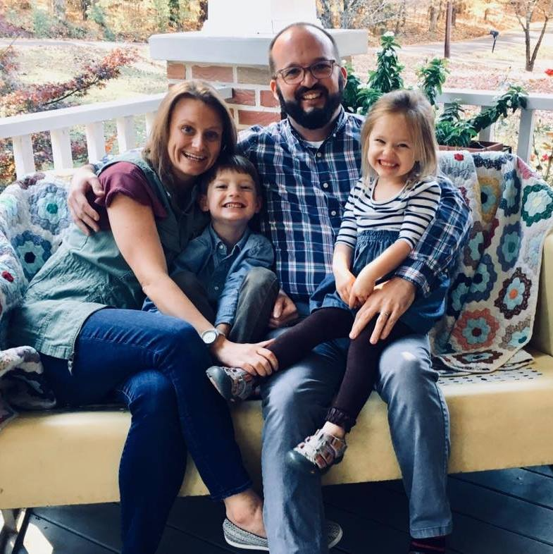 A family of four sits together on a couch, smiling, with a dog resting comfortably beside them.