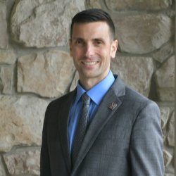 A man in a suit and tie stands confidently in front of a textured stone wall.