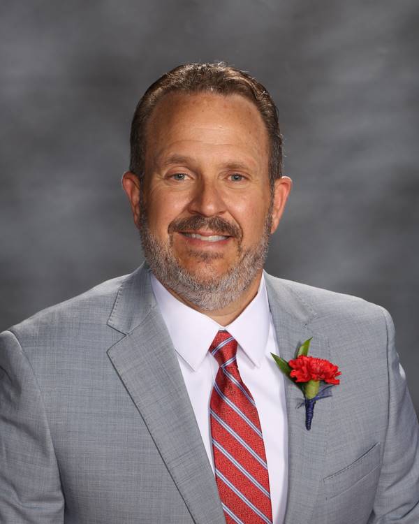 A man in a gray suit and red striped tie poses for a formal portrait, featuring a red carnation pinned to his lapel.
