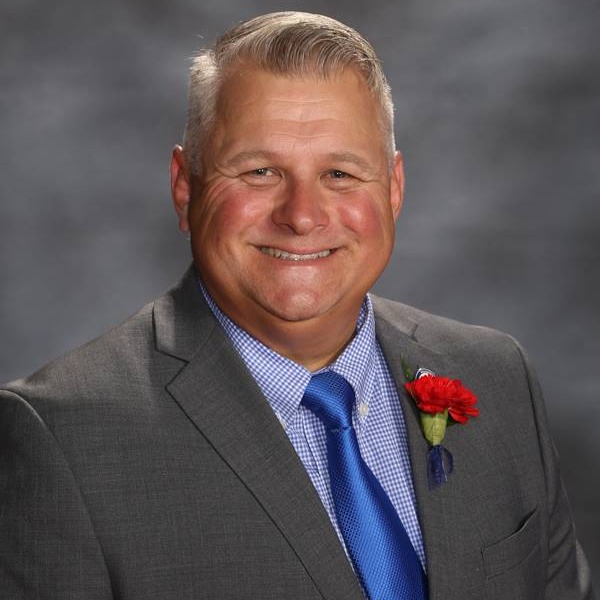 A man in a gray suit with a blue tie and a red boutonniere poses against a blurred gray background.