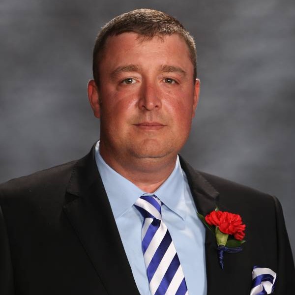 A man in a formal black suit with a light blue shirt and striped tie, adorned with a red flower boutonniere.