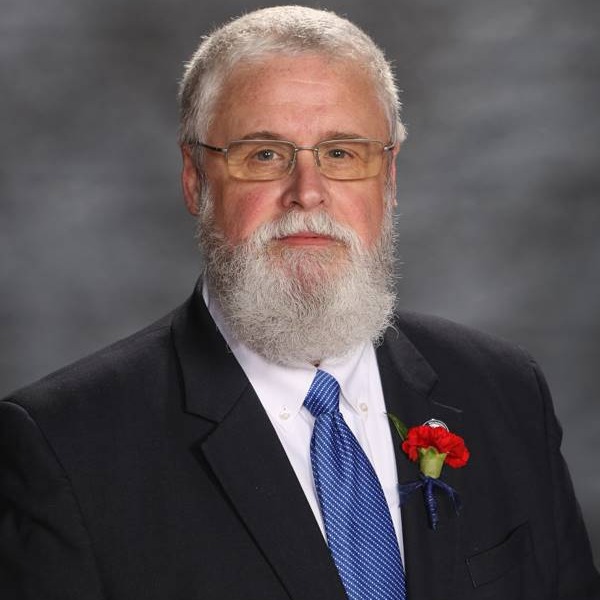 A distinguished man in a black suit and blue tie, adorned with a red flower pinned to his lapel, poses against a gray backdrop.