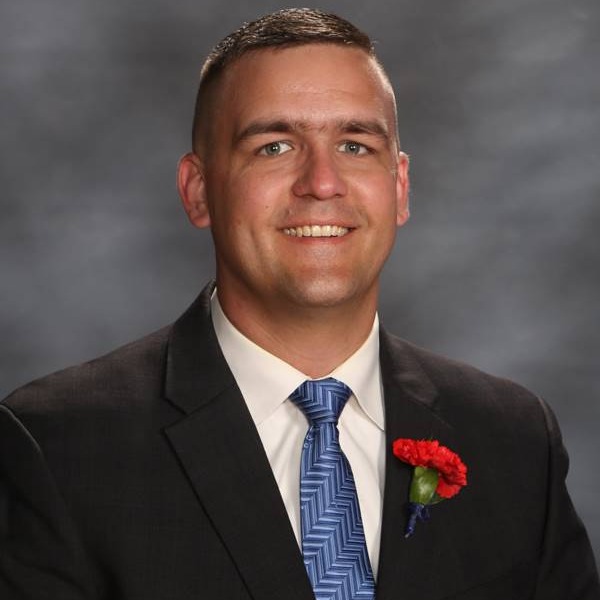 A man in a black suit with a blue tie and a red carnation boutonnière poses against a gray backdrop.