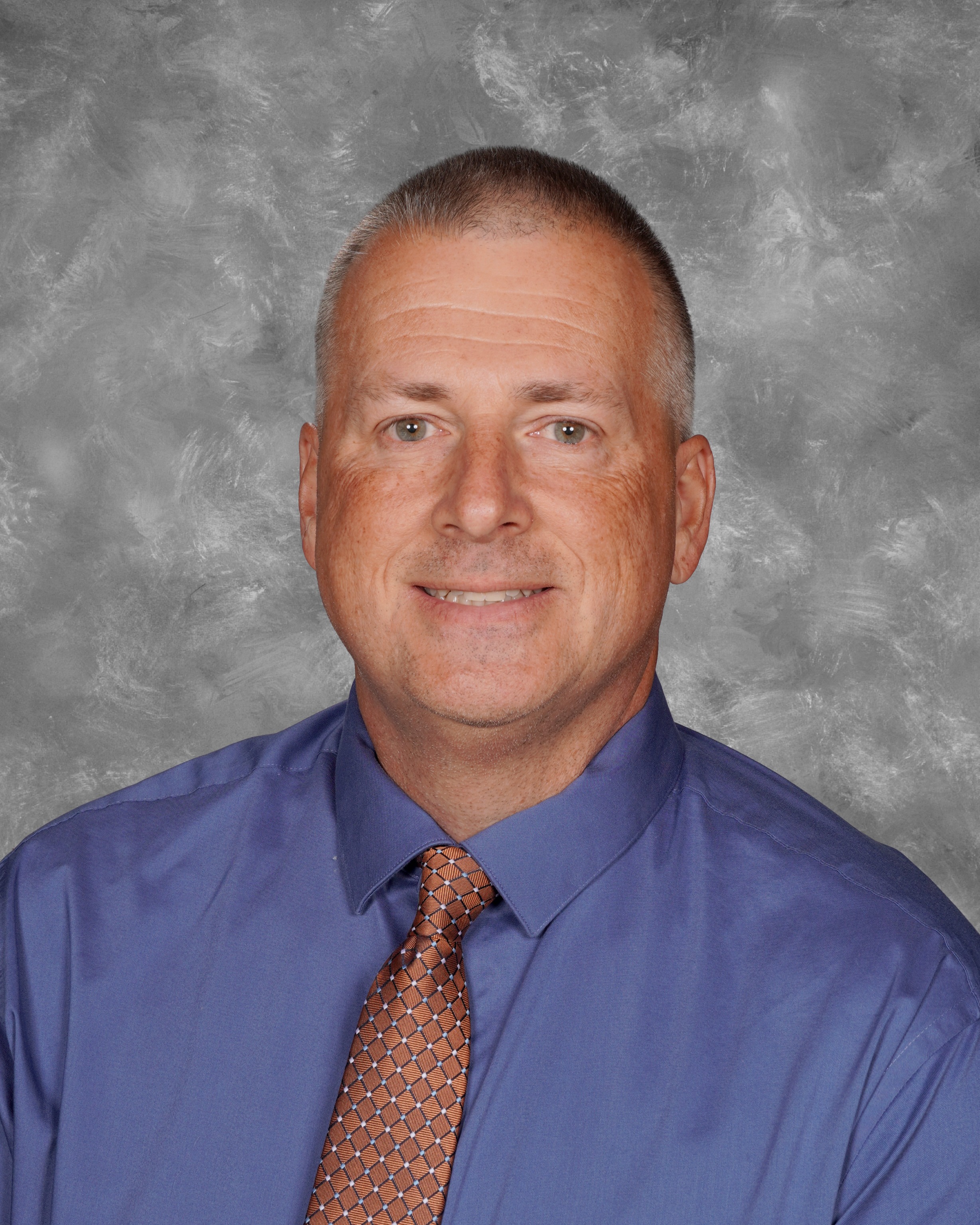 Bob Harcula school picture on a gray background. Bob is smiling, has a blue button up shirt on with a brown designed tie. 