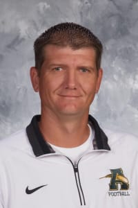 A man with short, spiky hair wearing a white athletic jacket with a black collar and a logo that includes the word 'Football.'