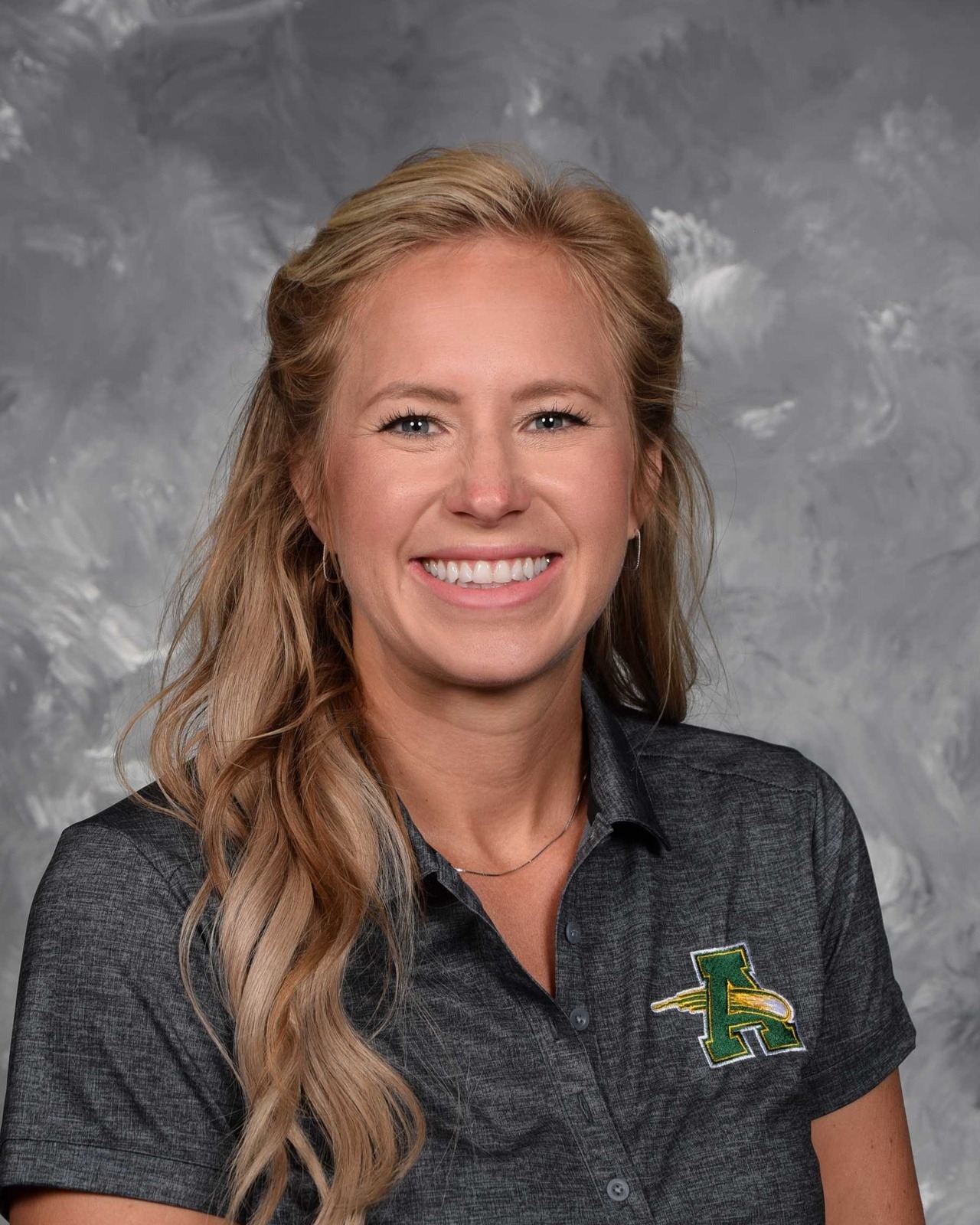 Professional head-and-shoulders portrait of a smiling person with long, wavy blonde hair styled half up and half down. She is wearing small hoop earrings, a silver necklace, and a dark gray collared polo shirt embroidered with a green and gold letter "A" logo on the left chest. The background is a mottled gray studio backdrop, and the subject is facing the camera with a friendly, approachable expression.