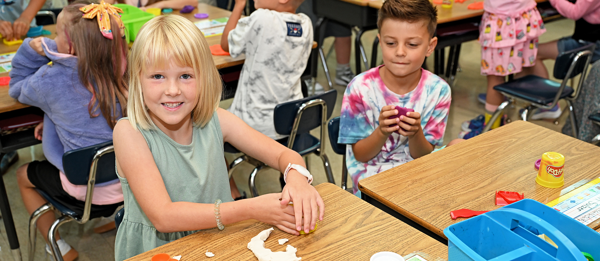 Children seated at desks in a classroom, one with blond hair crafting with clay.