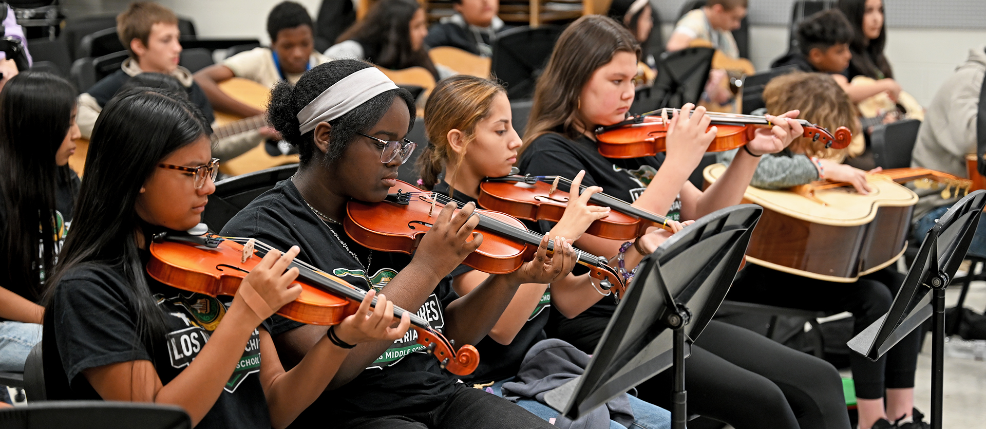 A classroom of students practicing violins and guitars. Some wear black shirts and some wear glasses.