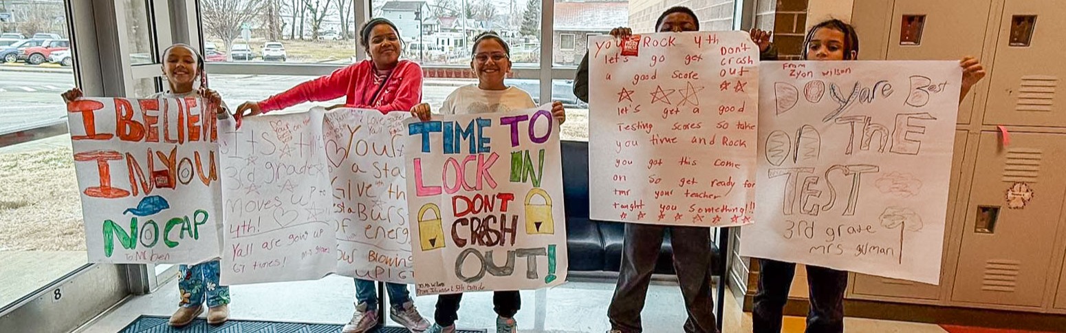 Students at Mary C. Snow West Side Elementary School standing at the entrance of the school holding encouraging signs for students to do well on their upcoming math test.