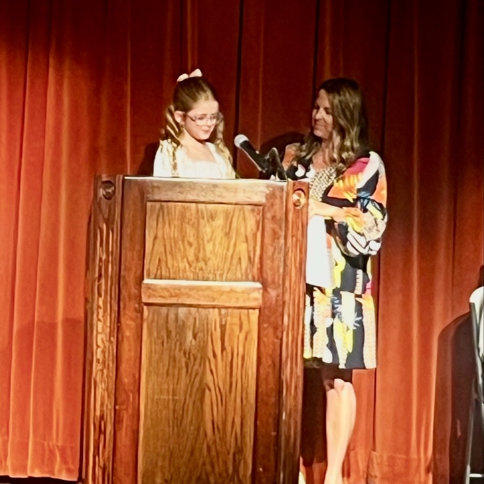 A student is standing at a podium on the SCHS auditorium stage reading a poem she wrote. A KCS employee is standing to the student's left watching her read.