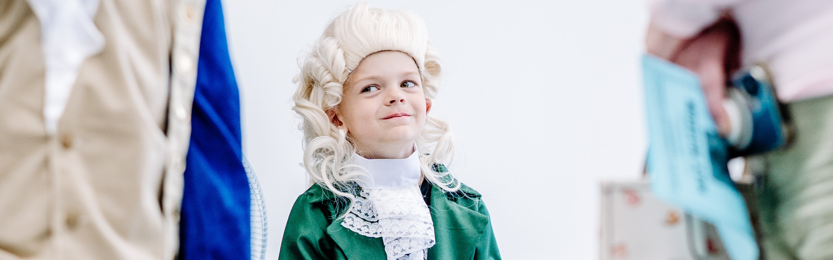 A student is dressed up as Thomas Jefferson for a school project. He is sitting in his classroom on a chair and smiling as he looks off to his right. 