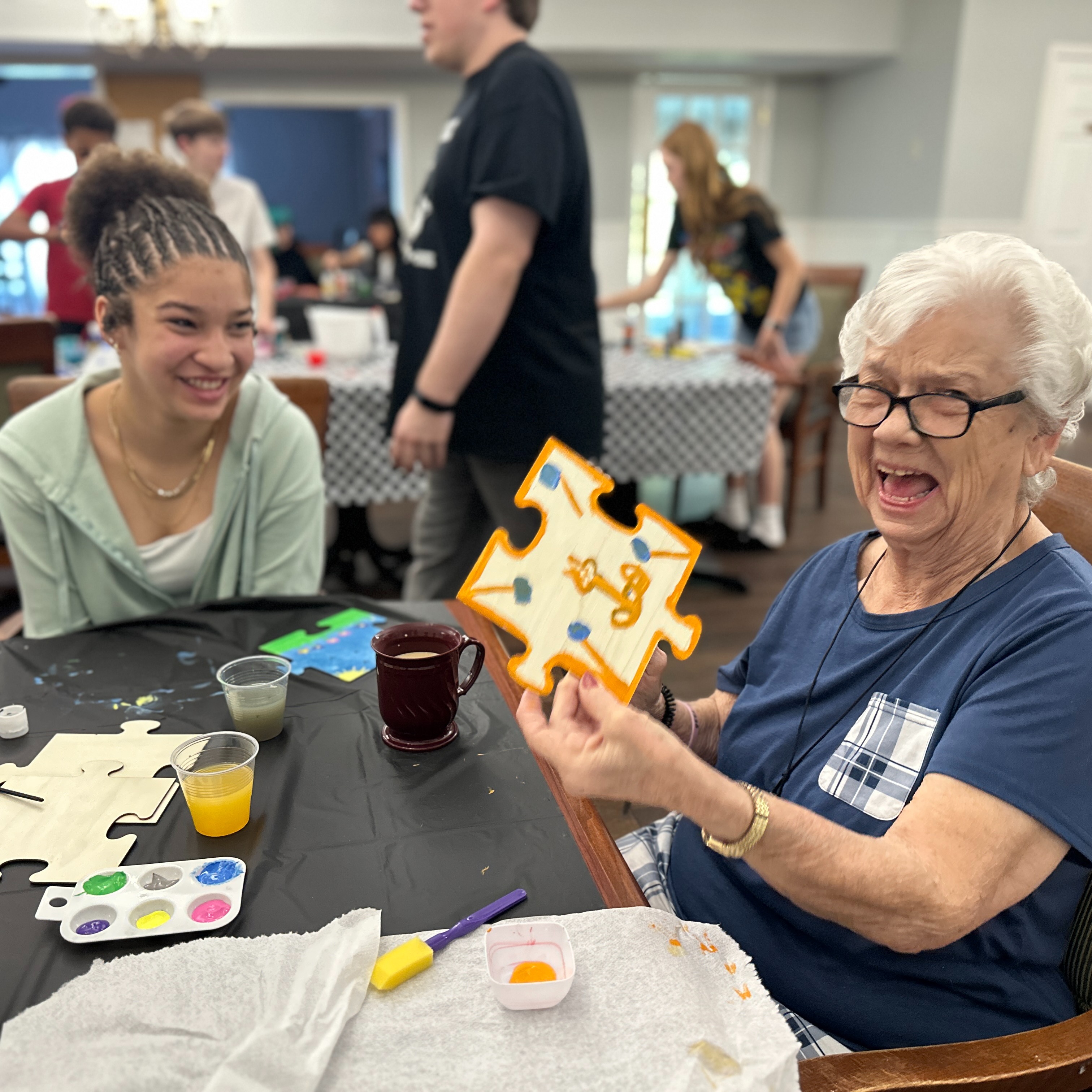 A nursing home resident, sitting at a table, is holding up a painting project she did with a student from Dunbar Middle School. The nursing home resident is laughing and the student is looking at the woman and smiling. 