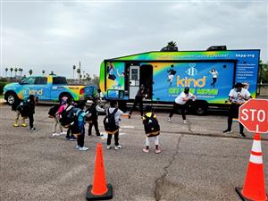 A group of children in backpacks gather near a colorful mobile unit promoting kindness, with traffic cones and a stop sign visible in the foreground.