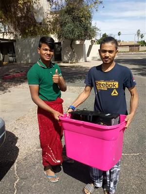 Two young men are standing outdoors, one giving a thumbs up while the other holds a large pink container filled with items, with a residential area in the background.