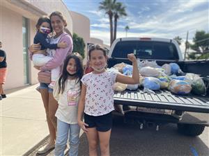 A group of four children and a woman stand in front of a pickup truck filled with bags of food, with palm trees and a blue sky in the background.
