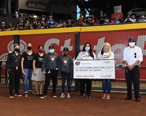 A group of individuals stands on a baseball field, holding a large check for a donation, with spectators visible in the background.
