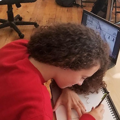 Student leans over her sketchbook as a computer sits open at her side