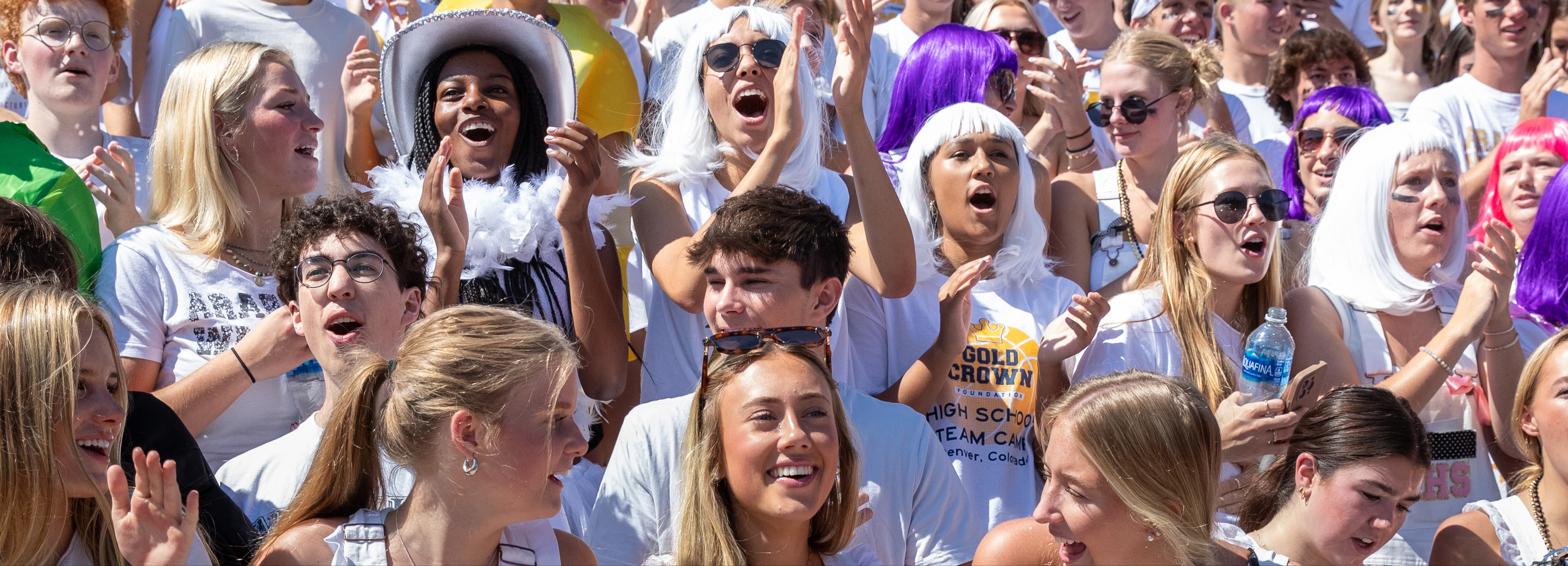 A large group of high school students cheering in an outdoor stadium. Many are wearing themed outfits and accessories, such as wigs, sunglasses, and feather boas. One student in the center wears a wide-brimmed hat and feather boa. Some have face paint or black marks under their eyes, and several hold water bottles. The students appear joyful and enthusiastic, showing strong school spirit and unity.