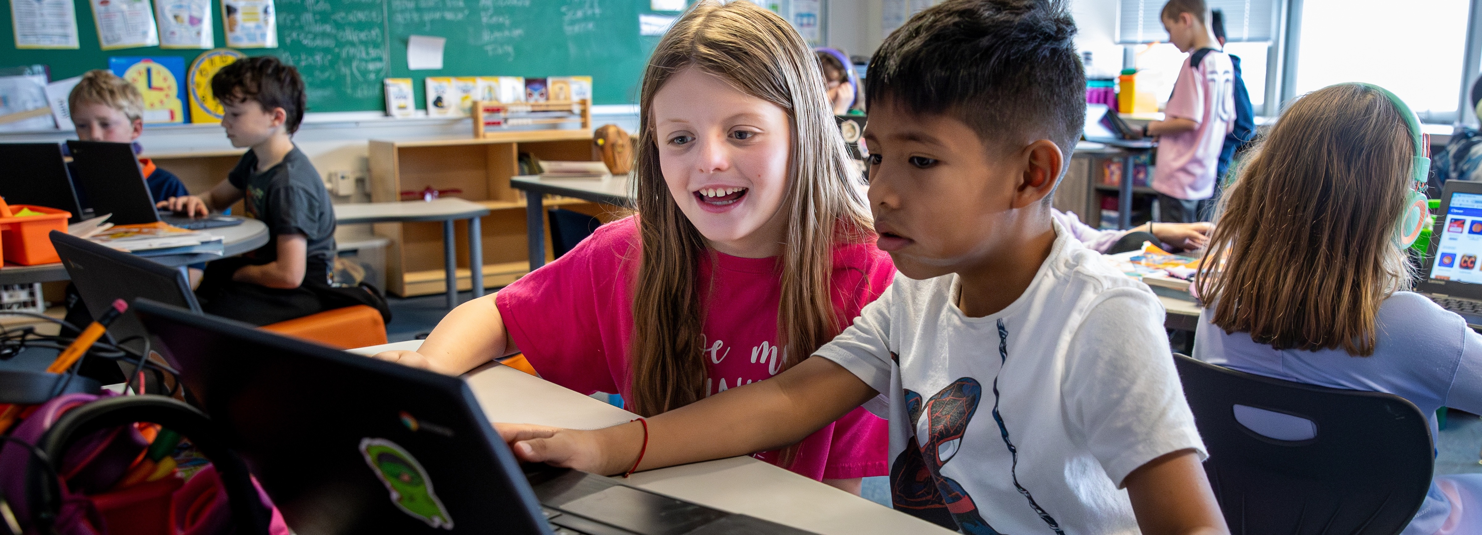Elementary-aged students working at laptops in a classroom. Two students at the front, a girl and a boy, are seated side by side and focused on the same screen, smiling and engaged. Other students are visible in the background at separate desks, some wearing headphones and using devices. The classroom has a chalkboard, books, educational posters, and a generally collaborative and tech-integrated environment.
