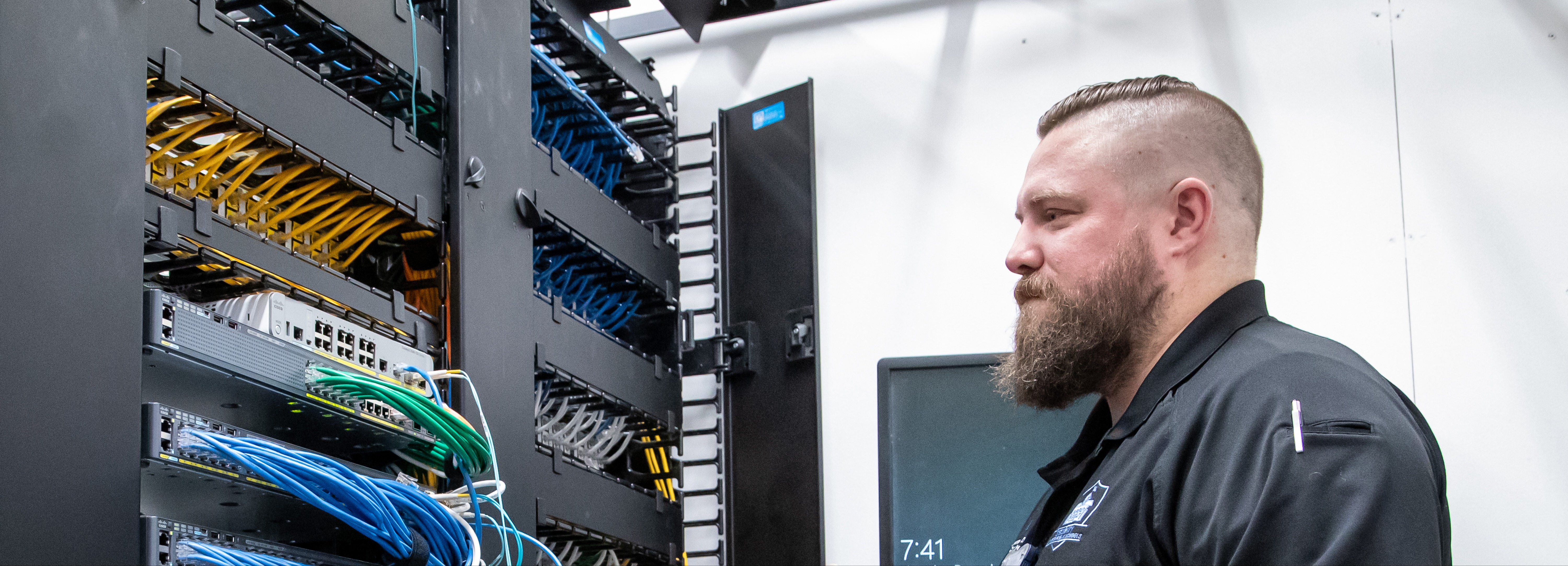An adult staff member standing in a server room, closely inspecting a tall rack of networking equipment. The equipment includes organized rows of cables and multiple hardware units with indicator lights. The staff member wears a collared shirt with an embroidered badge and has a neatly groomed beard and side-parted hair. A wall-mounted digital display in the background shows the time. The setting reflects a professional, behind-the-scenes view of IT operations within a school or district environment.