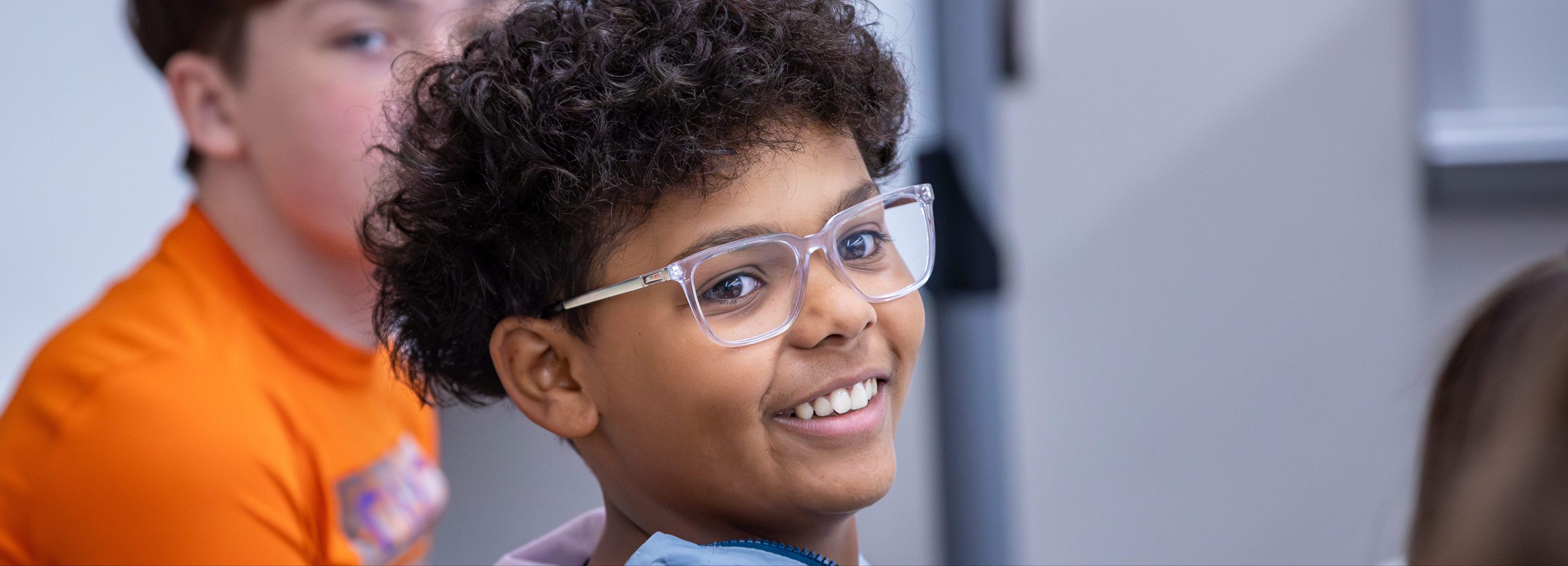Close-up of a smiling student wearing glasses, seated in a classroom. The student is looking slightly off-camera with a cheerful expression. Other students are partially visible in the background, creating a sense of a collaborative learning environment. The focus is on the student’s positive engagement and presence in the classroom.