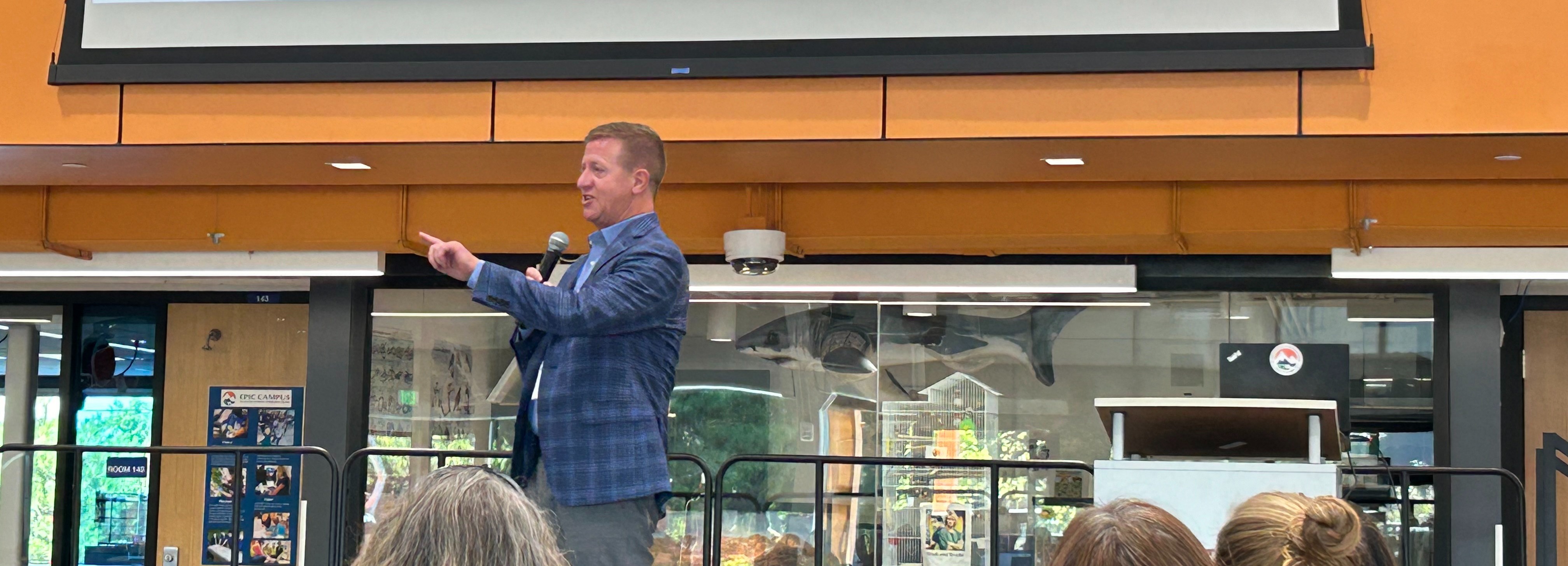 A school superintendent stands on a platform, speaking into a microphone and gesturing toward the audience. He wears a jacket and addresses a seated group of attendees, some of whom are partially visible in the foreground. Behind him are glass walls, a mounted shark model, and a doorway with posters. A blank projection screen hangs above the speaker, suggesting a presentation or assembly setting.